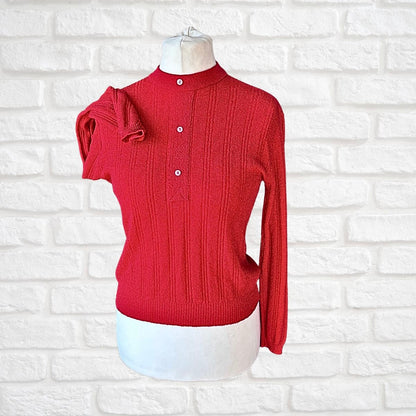 A red ribbed knit sweater displayed on a white dress form mannequin against a white painted brick wall. The sweater features a mock neck, a buttoned placket with white buttons, and a unique draped sleeve detail on the left side.