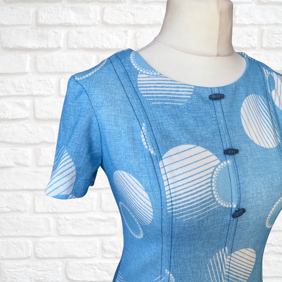 Blue dress with white abstract patterns on a mannequin against a white brick wall.