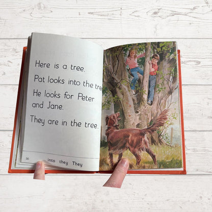 Open book with a story about children looking for someone in a tree, held by a hand on a light wooden surface.