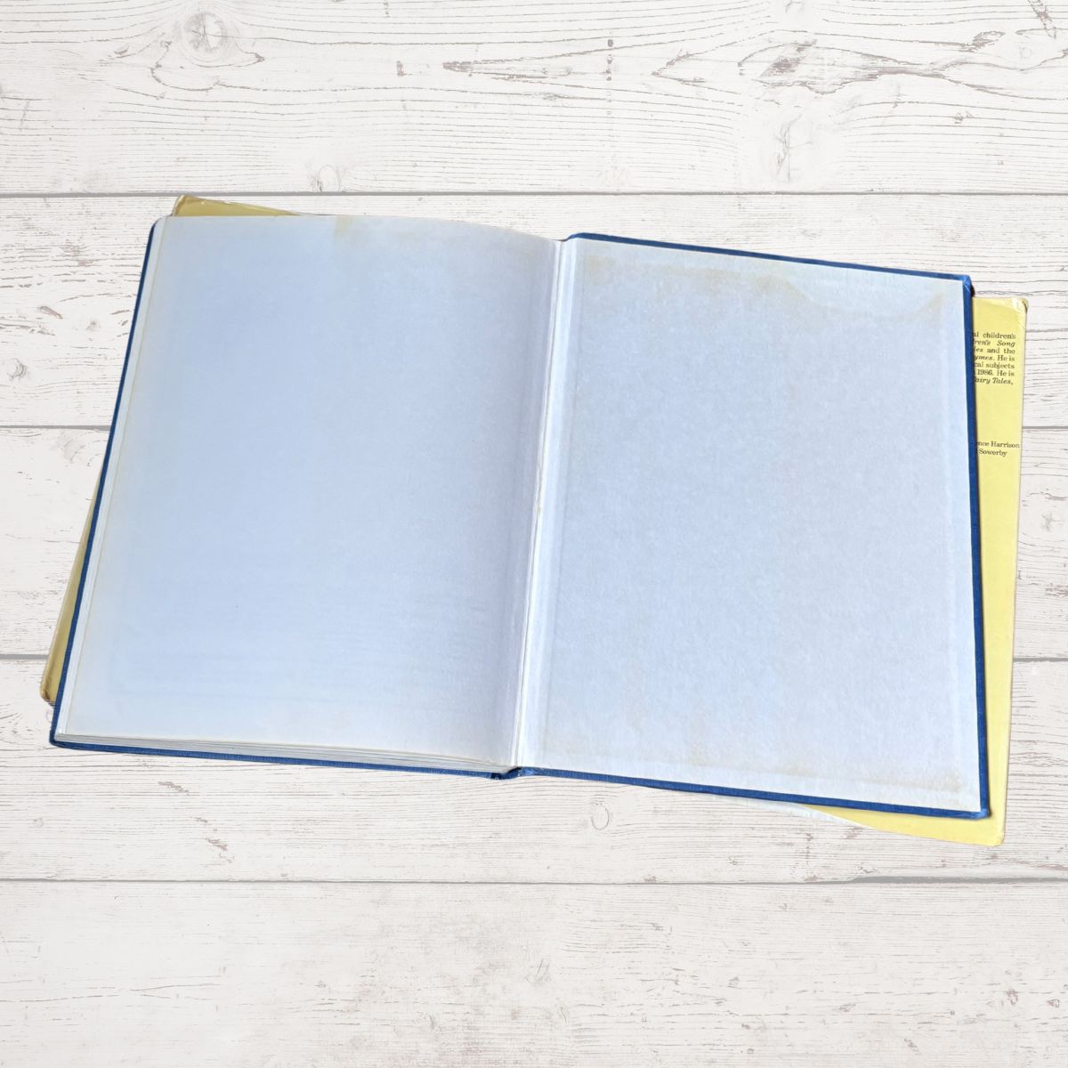 An open book with blank white pages, a blue cover edge, and a yellow interior page, placed on a light whitewashed wood surface.