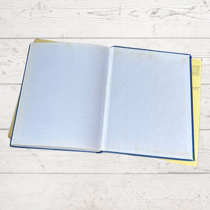 An open book with blank white pages, a blue cover edge, and a yellow interior page, placed on a light whitewashed wood surface.
