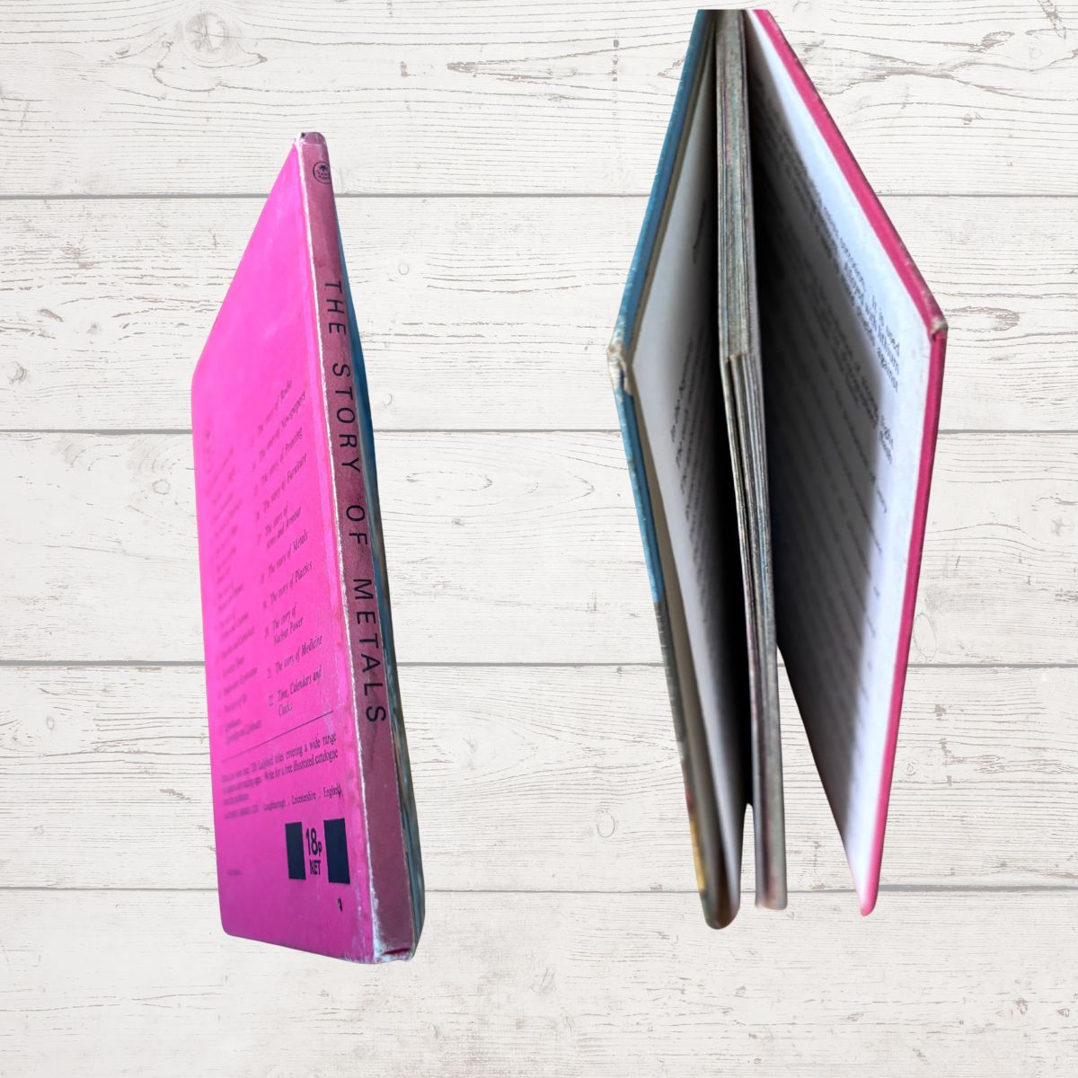 A vintage hardcover book titled 'THE STORY OF METALS' shown from two angles: standing upright to display its bright pink spine, and standing open to show the interior pages and blue/pink cover edges, set against a light wood plank background.