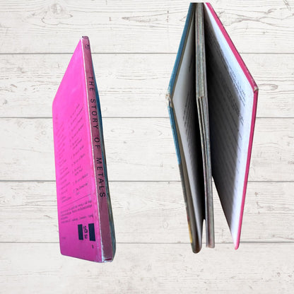 A vintage hardcover book titled 'THE STORY OF METALS' shown from two angles: standing upright to display its bright pink spine, and standing open to show the interior pages and blue/pink cover edges, set against a light wood plank background.
