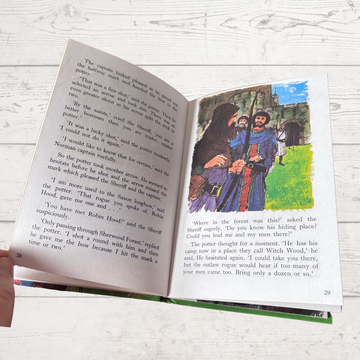 An open children's book lying on a white textured surface, showing printed text on both pages and a colorful illustration on the right page of two figures in a forest scene.