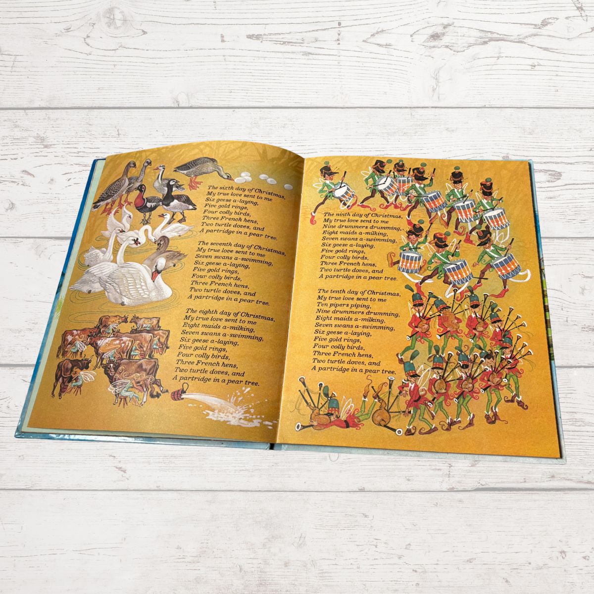 An open children's book spread on a white wood surface, showing illustrated Christmas scenes and printed text on both pages.