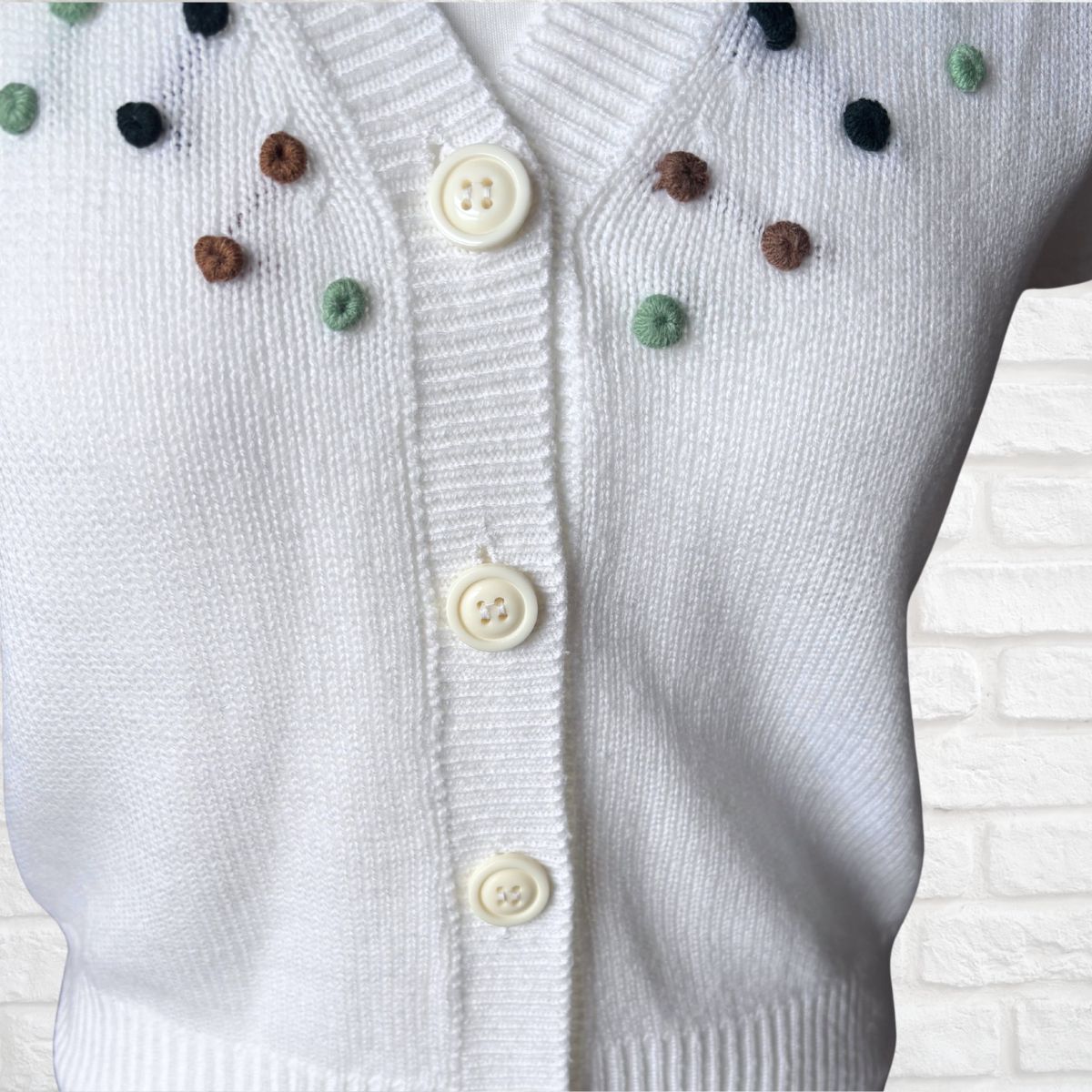 A close-up of a white knit cardigan sweater with cream buttons and small multicolored pom-pom accents on the chest, shown against a white brick wall background.
