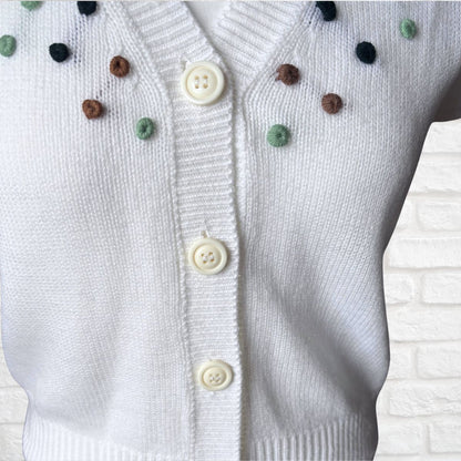 A close-up of a white knit cardigan sweater with cream buttons and small multicolored pom-pom accents on the chest, shown against a white brick wall background.