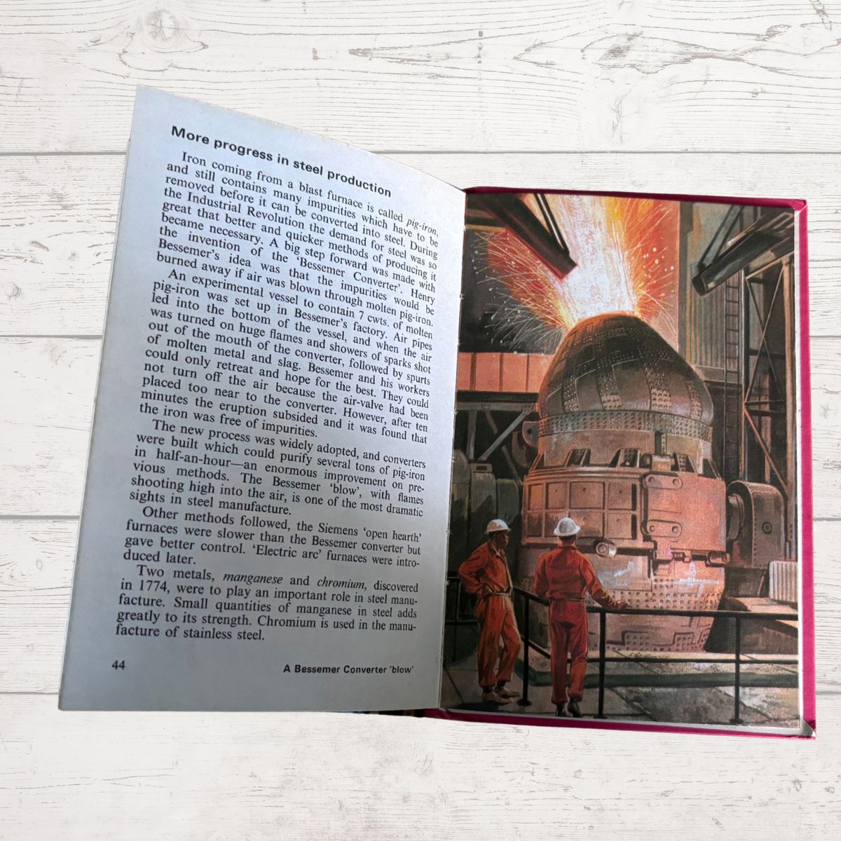 An open vintage book showing a page with text and a large illustration of workers in red overalls standing in front of a steel furnace, photographed on a white wood surface.