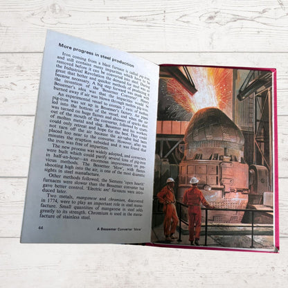 An open vintage book showing a page with text and a large illustration of workers in red overalls standing in front of a steel furnace, photographed on a white wood surface.