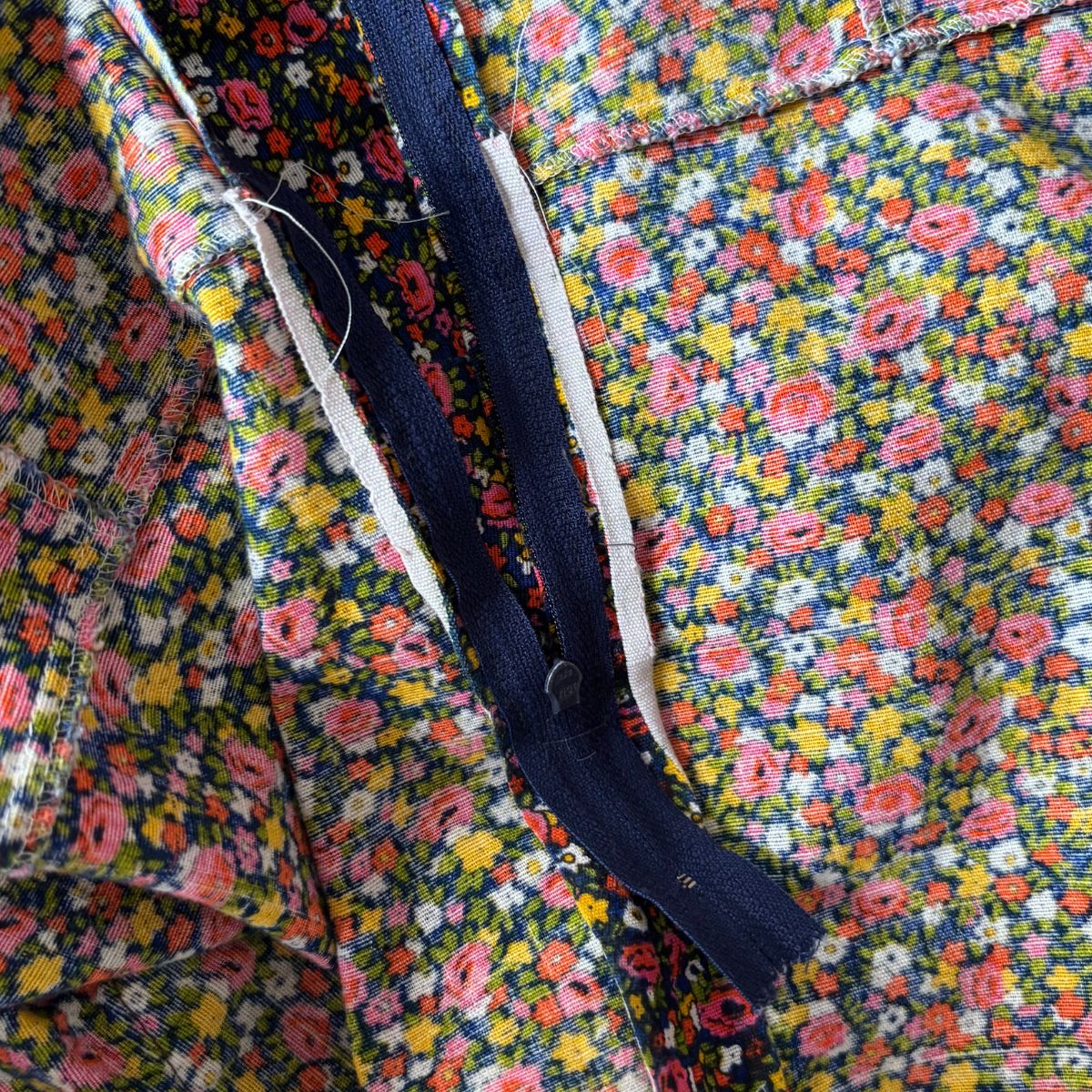 Close-up of a floral fabric garment or textile with a dense multicolor print, a dark navy zipper, and white trim along the zipper.