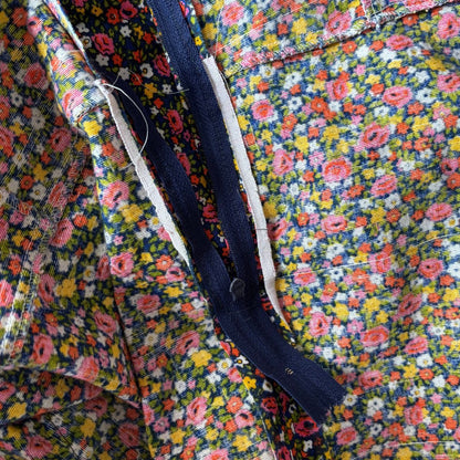 Close-up of a floral fabric garment or textile with a dense multicolor print, a dark navy zipper, and white trim along the zipper.
