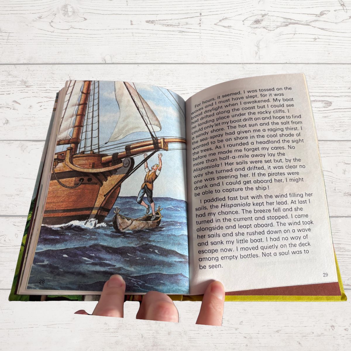 A children's picture book being held open by two hands, showing a colorful illustration of a sailing ship on one page and printed text on the other, set against a light wood background.