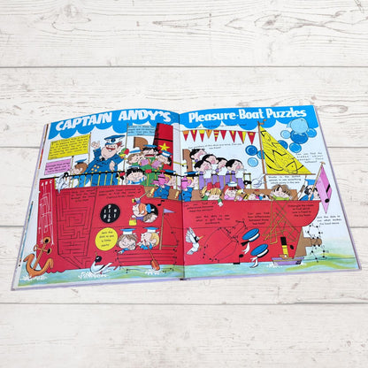 An open children's activity book titled 'Captain Andy's Pleasure Boat Puzzles' laid flat on a white wood background. The pages feature colorful cartoon illustrations of Captain Andy and characters on a red boat, with puzzle sections and text boxes.