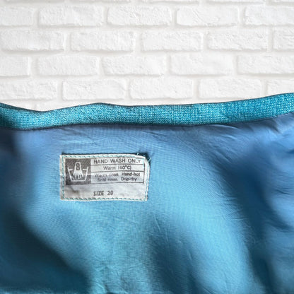 A close-up of a blue fabric item, likely a towel or blanket, showing a white sewn-in label that reads 'HAND WASH ONLY' and 'SIZE 28'. The fabric has a textured, glittery teal-blue top edge and a smooth, slightly crinkled blue body, set against a white brick wall background.