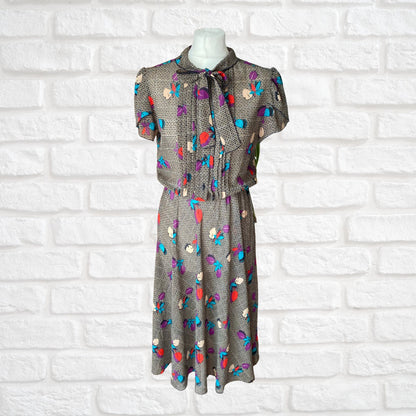 A vintage botanical print dress with a pleated bodice, black buttons, and a pussy bow collar, displayed on a mannequin against a white brick wall background.