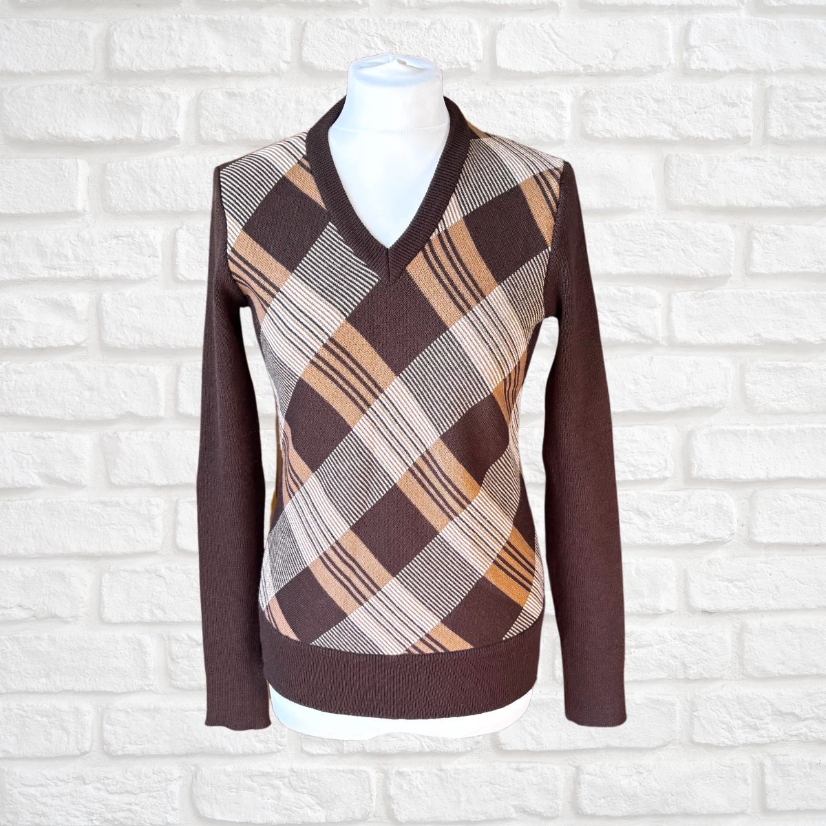 A brown long-sleeve V-neck sweater displayed on a white mannequin torso against a white brick wall background. The sweater features a brown, beige, and white plaid pattern across the chest and shoulders.