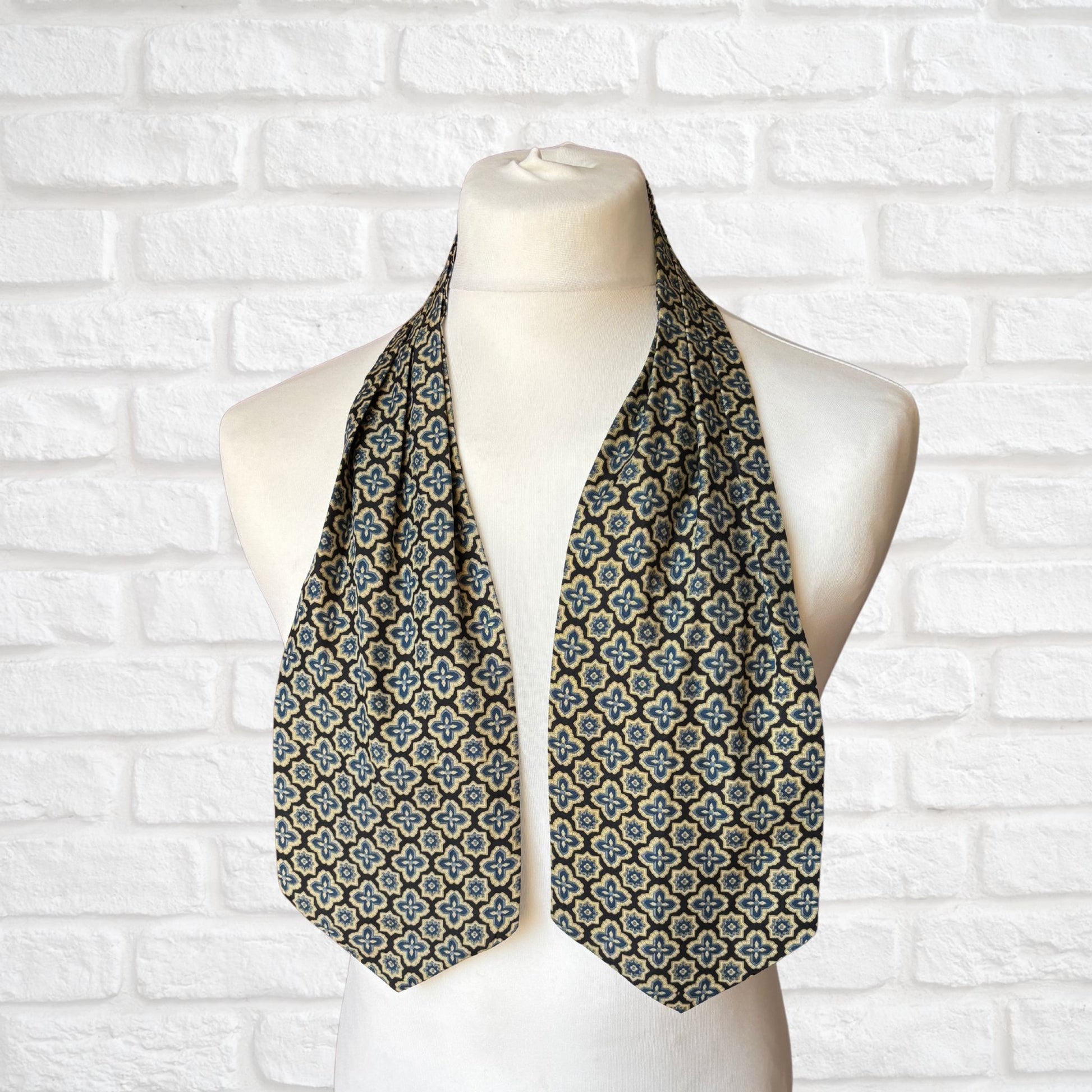 A vintage Austin Reed cravat featuring a black necktie with a cream, gold, and blue stylized floral print, displayed on a mannequin against a white brick wall background.