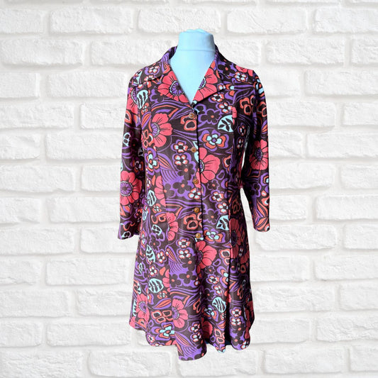A long-sleeve collared button-front dress displayed on a light blue mannequin torso against a white painted brick wall. The dress has a bold retro floral and paisley print in purple, pink, orange, and teal.