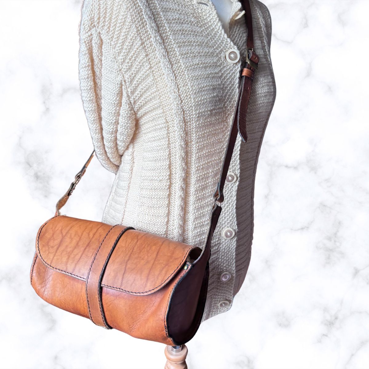 A brown leather crossbody bag with a curved flap and dark brown trim, shown on a cream-colored knit dress form against a white marble background.