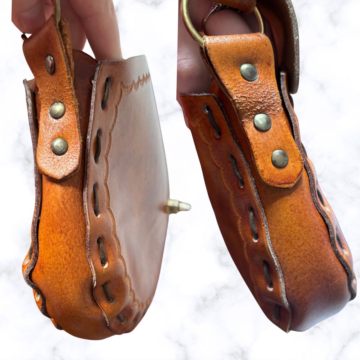 Leather shoulder bag with brass rivets and a metal ring, held up by a hand against a white marble background