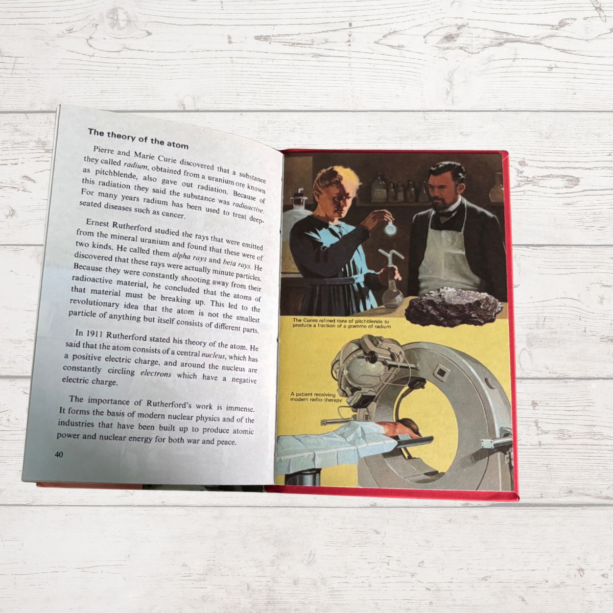 An open vintage book  showing printed pages about the history of the atom, photographed on a white wood surface. The left page is mostly blank with text, and the right page contains a photo of a man holding a model atom with a red border.