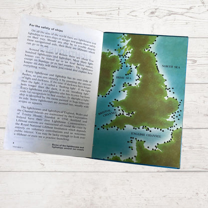 An open children's book spread showing a page with text about lighthouses and a map of the British Isles with lighthouses marked in green dots, placed on a light wood surface.