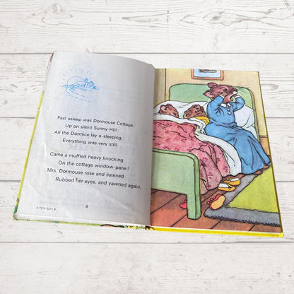 An open children's book lying on a white wood surface, showing a printed illustration of a woman in a blue dress waking up from a sleeping bed in a cottage scene.