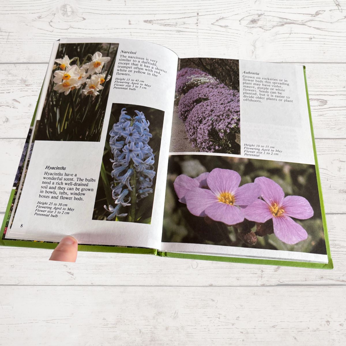 An open book showing printed pages with flower photographs and text, held by a visible finger, photographed on a light wood surface.