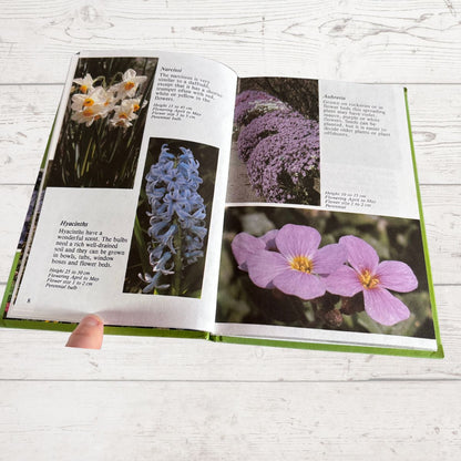 An open book showing printed pages with flower photographs and text, held by a visible finger, photographed on a light wood surface.