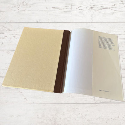 An open book lying on a white wood plank surface, with a textured beige cover on the left, a dark brown spine band, blank white pages in the center, and printed text on the right page.