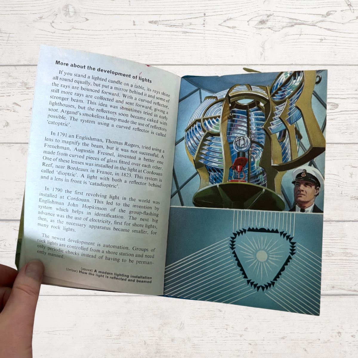 An open book showing printed text on the left page and a large illustrated maritime scene with a lighthouse and a man on the right page. A hand is holding the lower left corner of the book against a white wood background.