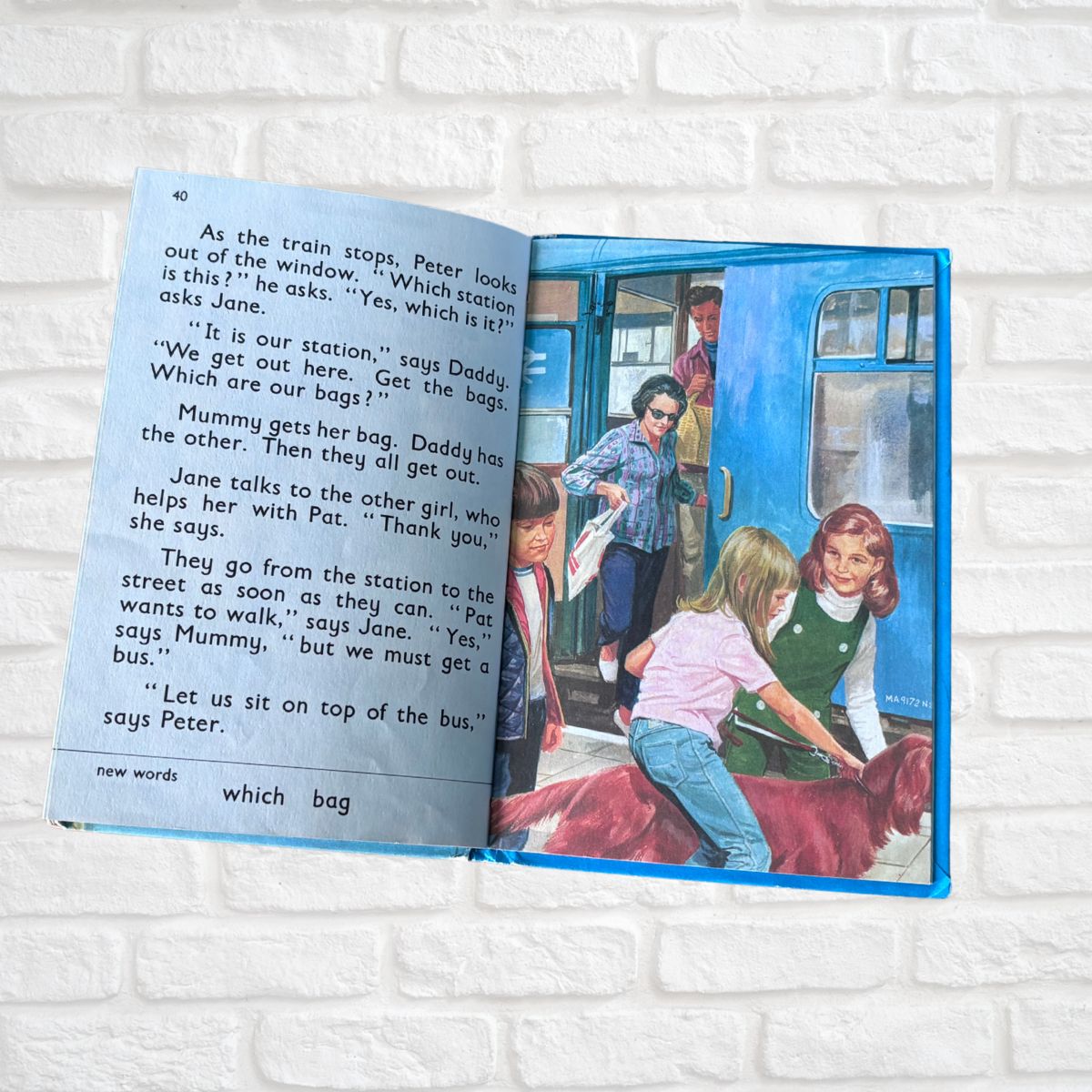 Open book with a colorful illustration of children at a train station on a white brick background.