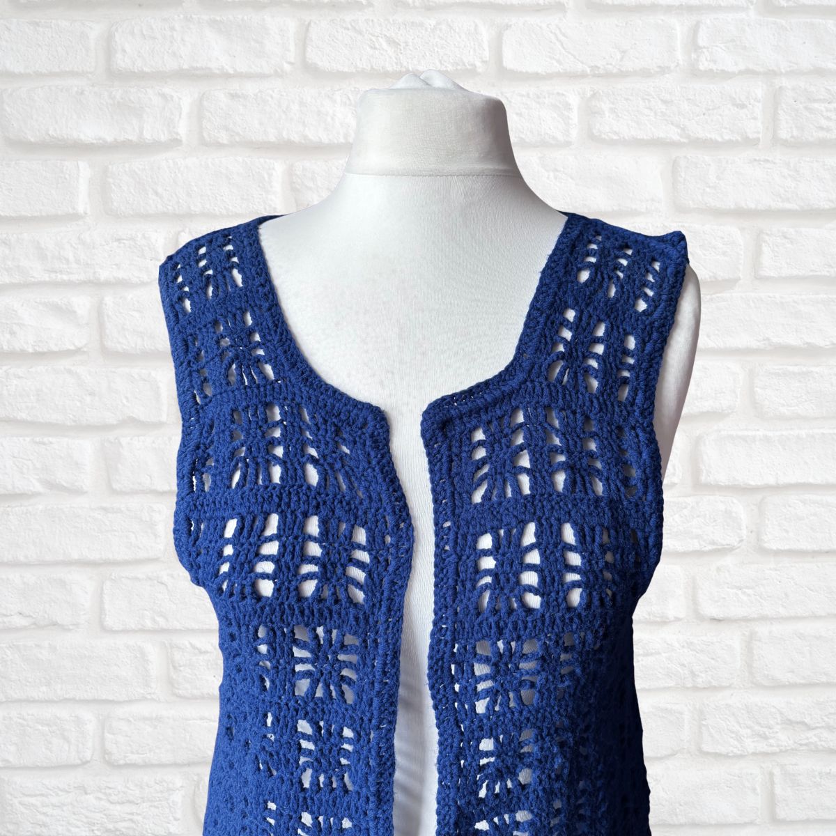 A blue open-front crochet vest displayed on a white mannequin torso against a white brick wall background.