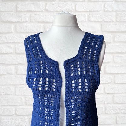 A blue open-front crochet vest displayed on a white mannequin torso against a white brick wall background.