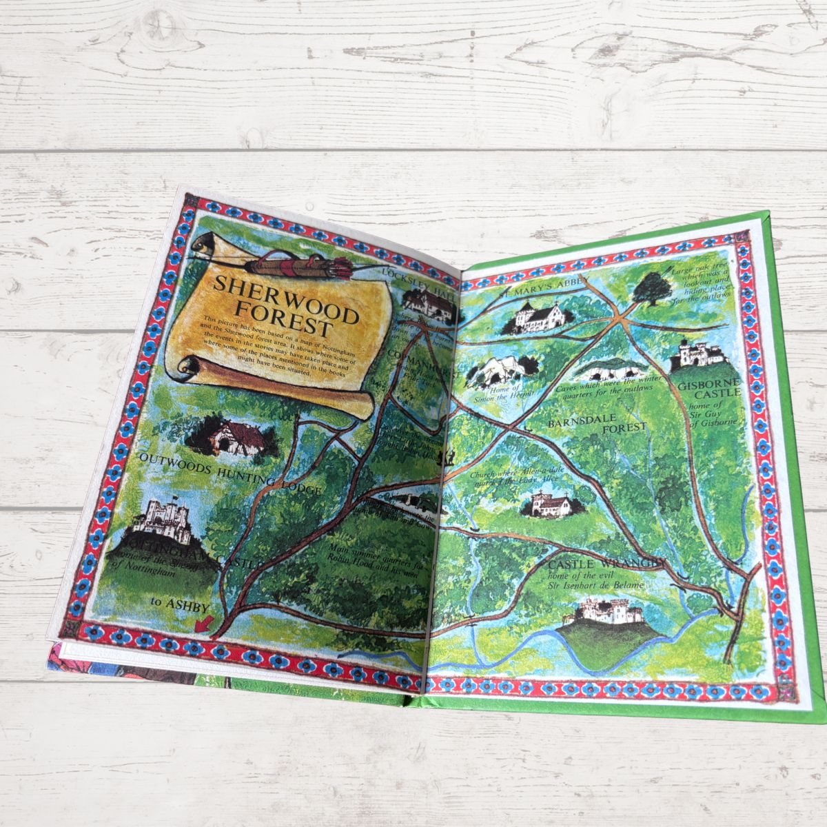 An open illustrated map book titled 'Sherwood Forest' laid flat on a light whitewashed wood surface. The pages show a colorful fantasy-style forest map with trees, paths, houses, and castle illustrations.