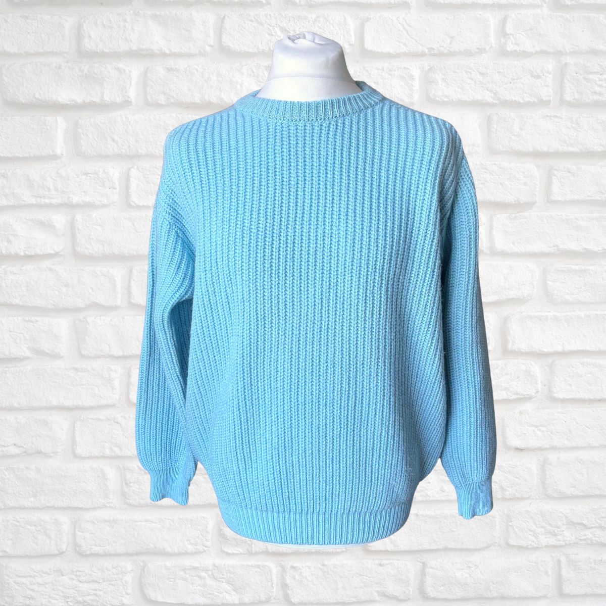 A light blue ribbed knit sweater displayed on a white mannequin torso against a white painted brick wall background.