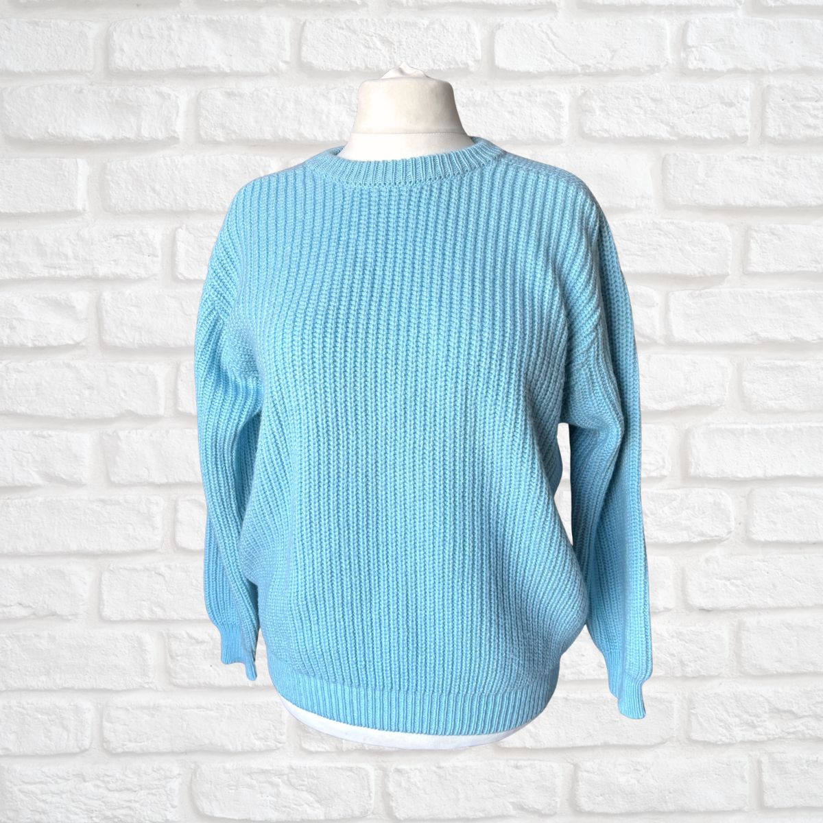 A light blue ribbed knit sweater displayed on a white mannequin torso against a white painted brick wall background.
