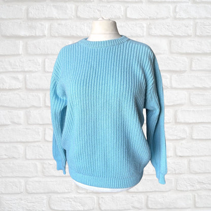 A light blue ribbed knit sweater displayed on a white mannequin torso against a white painted brick wall background.