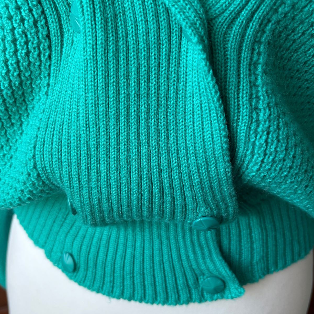 Close-up of a teal green ribbed knit cardigan sweater on a white mannequin torso, showing the buttoned front and knit texture.