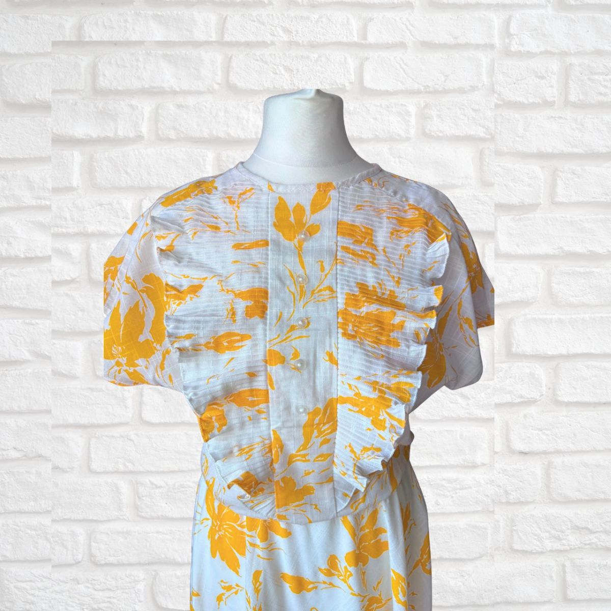 White dress with yellow floral pattern on a mannequin against a white brick wall.