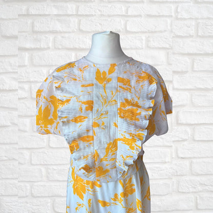 White dress with yellow floral pattern on a mannequin against a white brick wall.