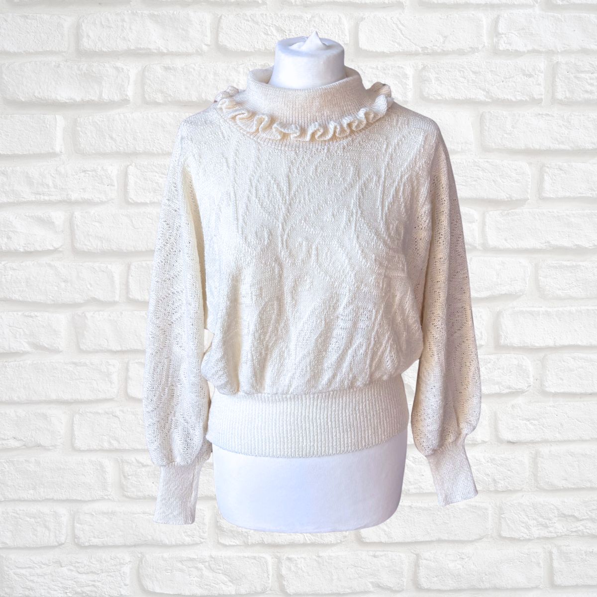 An off-white cable-knit sweater with a ruffled mock neck collar, displayed on a white mannequin torso against a white painted brick wall background.