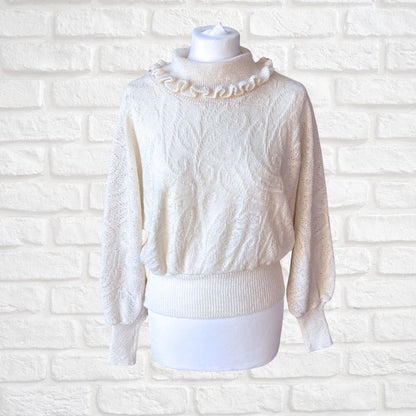 An off-white cable-knit sweater with a ruffled mock neck collar, displayed on a white mannequin torso against a white painted brick wall background.