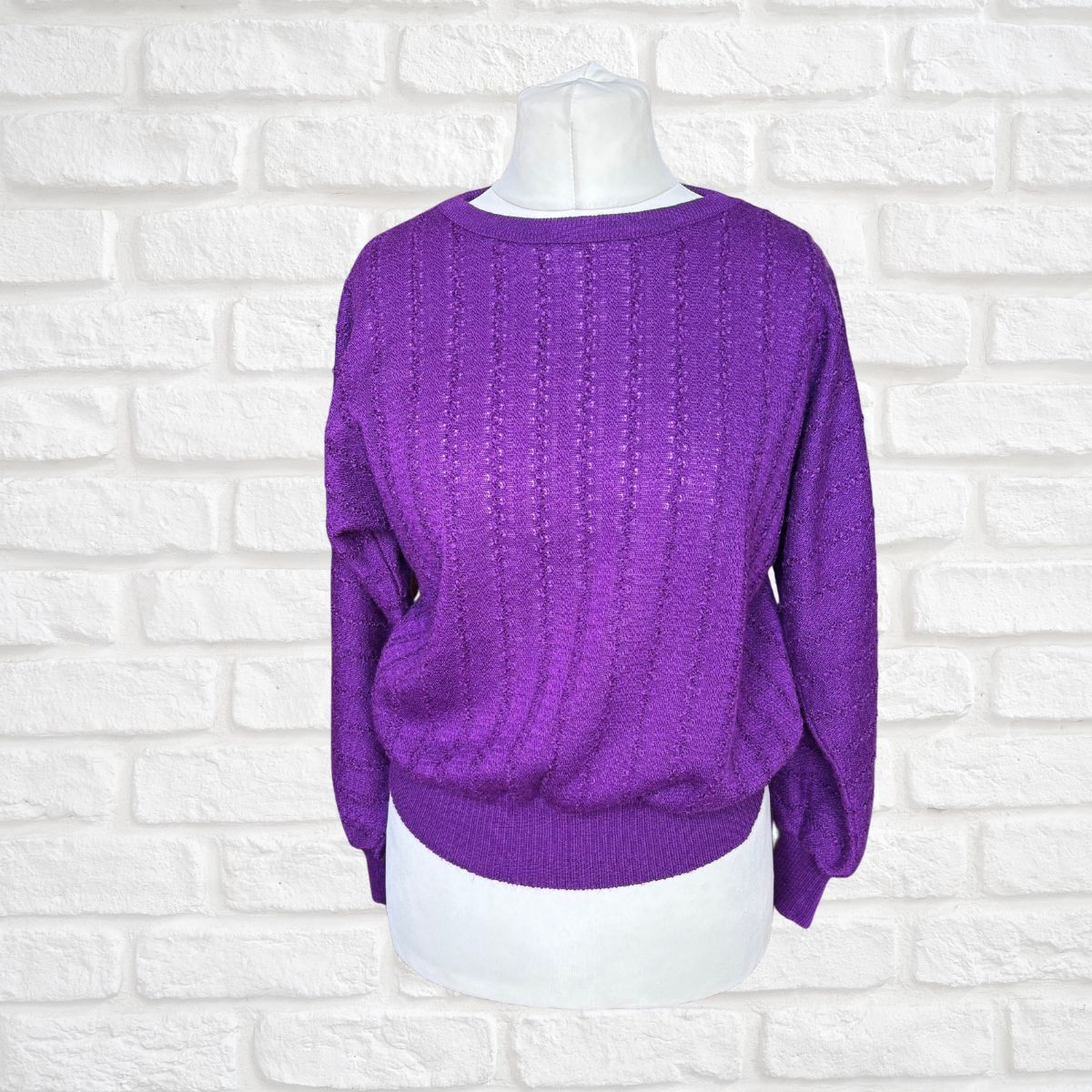 A purple textured knit sweater displayed on a white mannequin torso against a white brick wall background.