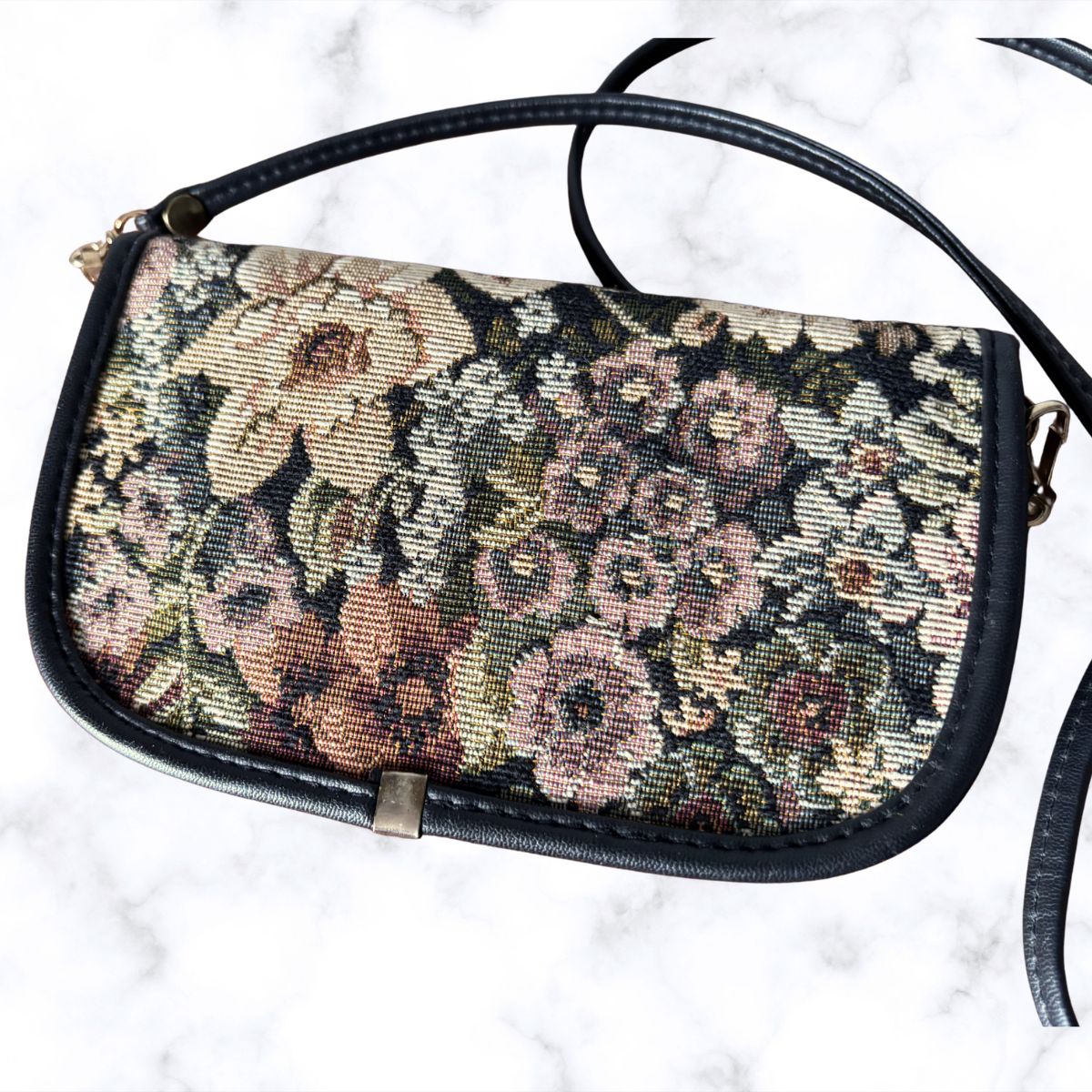 A vintage floral tapestry clutch bag with black leather trim and strap, displayed on a white marble surface.