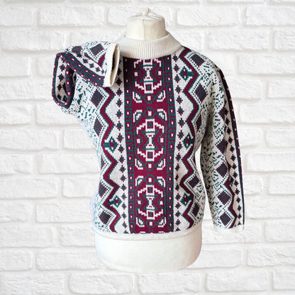 A patterned knit sweater displayed on a cream mannequin torso against a white painted brick wall. The sweater has a cream ribbed mock neck and cuffs, with a bold geometric Fair Isle style pattern featuring vertical bands of burgundy, cream, navy, and small teal accents.