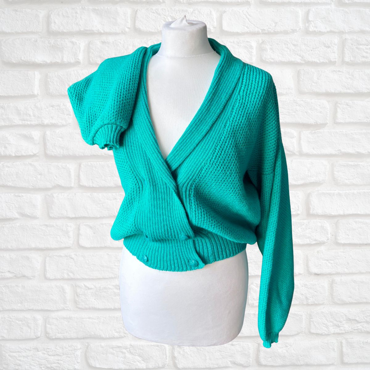 A bright turquoise knit wrap cardigan sweater displayed on a white mannequin torso against a white painted brick wall background.