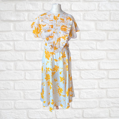White dress with yellow floral pattern on a mannequin against a white brick wall.