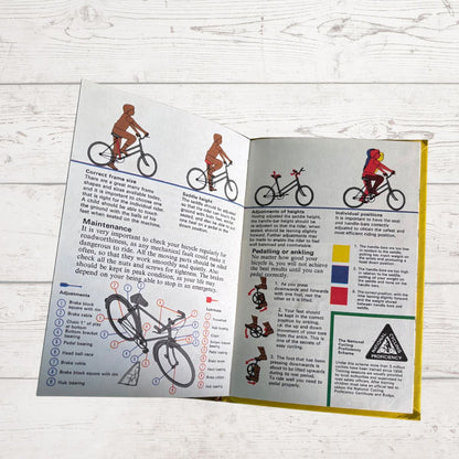 An open printed bicycle safety instruction booklet laid flat on a white wood surface. The booklet contains illustrated riders, bicycle diagrams, maintenance tips, and emergency braking instructions.