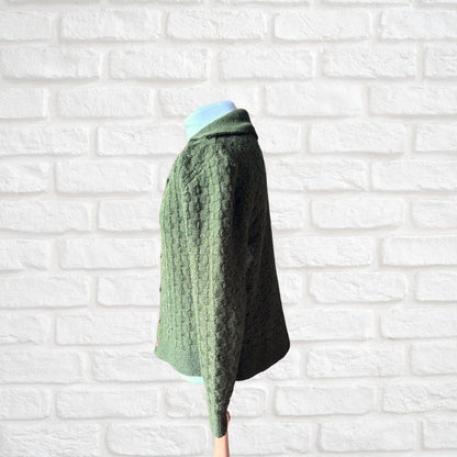 A sage green cable-knit cropped cardigan sweater displayed on a mannequin torso against a white painted brick wall background.